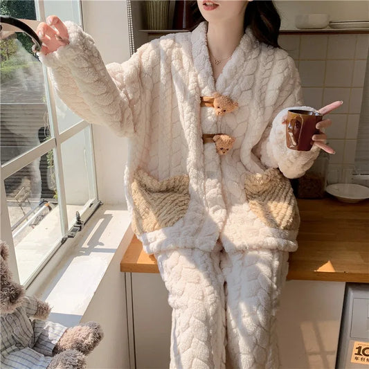 A woman wearing a cozy cream-colored quilted pajama set with beige patch pockets and bear-shaped toggle closures, standing in a kitchen and holding a coffee cup.