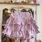 A light pink tiered ruffle skirt with lace-up front details and long ribbon ties, hanging on a wooden hanger in front of a white cabinet.