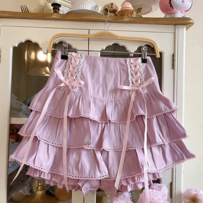 A light pink tiered ruffle skirt with lace-up front details and long ribbon ties, hanging on a wooden hanger in front of a white cabinet.