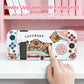 kawaii capybara watermelon switch case, this is the white version with three capybaras holding onto a watermelon while displaying different happy expressions on their faces, this image is also stating the the case is anti fingerprint, this is displayed on a white desk with a pink bookcase.