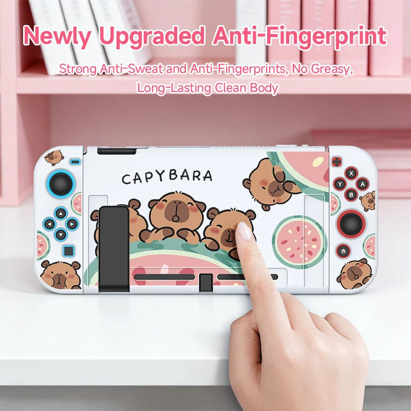 kawaii capybara watermelon switch case, this is the white version with three capybaras holding onto a watermelon while displaying different happy expressions on their faces, this image is also stating the the case is anti fingerprint, this is displayed on a white desk with a pink bookcase.