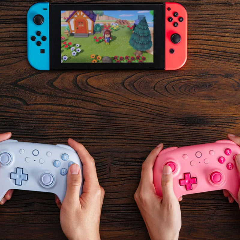 Two gaming controllers, one blue and one pink, being used on a wooden surface with a screen displaying a game.