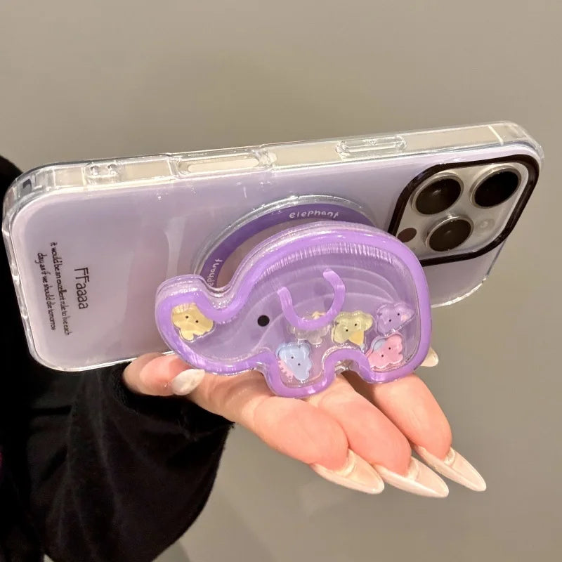 a woman holding a  phone with the kawaii cute elephant quicksand phone grip , this is in the color purple with colorful elephant faces inside, displaying it folds out and can be tilted to hold your phone on a flat surface, this is on a white background.