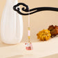 A pink and gold Japanese maneki-neko lucky cat charm hanging from a black decorative hair stick, set against a warm beige background with a white vase and dried flowers.