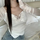 White lace top worn by a person with sheer sleeves, sitting on a bed.