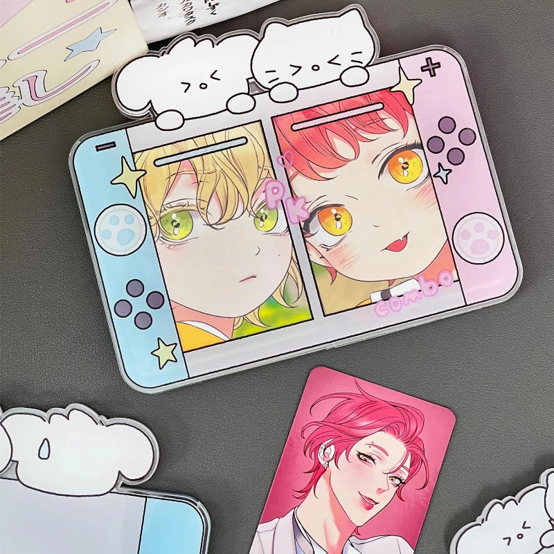 A novelty acrylic desk stand designed to look like a split Nintendo Switch console, featuring anime character illustrations on the blue and pink halves. The case is shown on a dark gray surface alongside other matching anime-themed accessories, including cloud-shaped stickers and a character card.