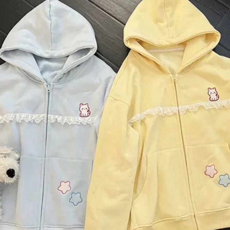 Two pastel zip-up hoodies laid flat on a dark wood floor: one light blue and one pale yellow, both with lace trim across the chest, small cat embroidery, star patches, and front pockets.