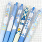 Five cute cartoon gel pens with blue and white patterns, including checkered designs, arranged diagonally on a white grid paper background.