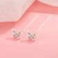 A pair of silver butterfly drop earrings with pink and yellow gemstone accents, displayed on a soft pink background with a sheer white fabric in the upper left corner.
