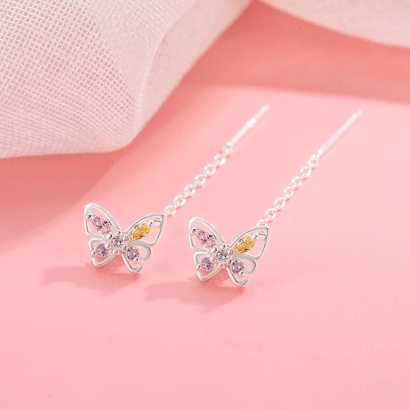 A pair of silver butterfly drop earrings with pink and yellow gemstone accents, displayed on a soft pink background with a sheer white fabric in the upper left corner.