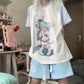 A person wearing a light blue and white raglan t-shirt with a large anime character graphic on the front, paired with matching light blue shorts in a bedroom setting.