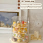 A three-tier rotating display stand with a clear cylindrical outer shell and cream-colored shelves, holding small pastel character figurines. The stand sits on a tabletop in a styled room setting with framed wall art, a small decorative object, and text callouts describing its 360° rotatable design, entry-level storage suitability, and three-layer cake shelf capacity.