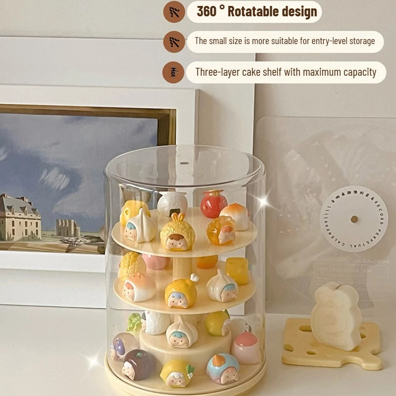 A three-tier rotating display stand with a clear cylindrical outer shell and cream-colored shelves, holding small pastel character figurines. The stand sits on a tabletop in a styled room setting with framed wall art, a small decorative object, and text callouts describing its 360° rotatable design, entry-level storage suitability, and three-layer cake shelf capacity.