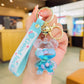 Hand holding a blue keychain with a transparent figure and 'Good Luck' tag in a blurred indoor setting.