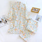 A two-piece pajama set laid flat on a white surface, featuring a short-sleeve top and matching shorts in an all-over pastel floral print. The set is styled with a wooden calendar, a magazine, and a pillow.