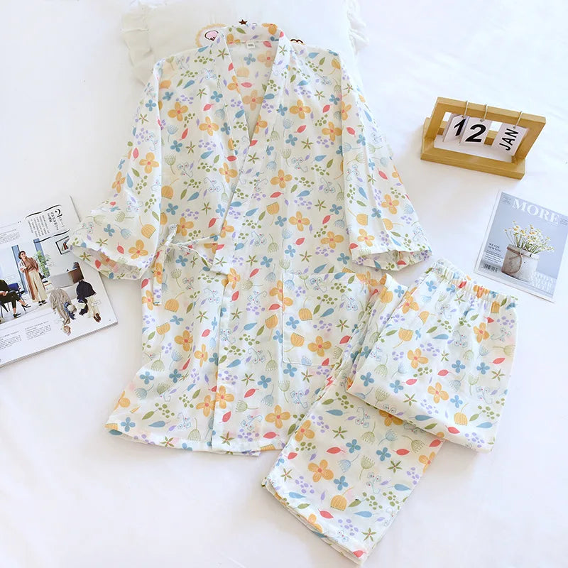 A two-piece pajama set laid flat on a white surface, featuring a short-sleeve top and matching shorts in an all-over pastel floral print. The set is styled with a wooden calendar, a magazine, and a pillow.