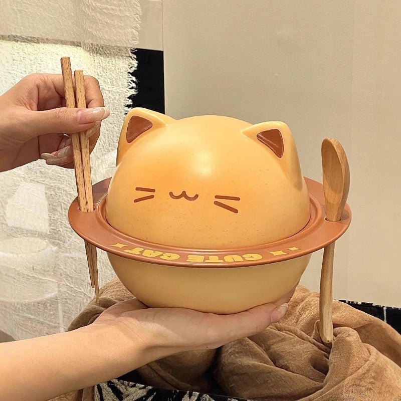 A cute, cat-shaped ceramic bowl with a lid, held by hands with wooden chopsticks and a spoon resting in its side holders. The bowl is a warm peachy-orange color with a brown face and features the text 'IT CUTE CAT!' on the rim.
