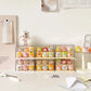 Six clear acrylic display boxes stacked horizontally on a tabletop, each holding multiple small pastel character figurines. The setup is styled in a clean, minimalist room with a cream wall, a small white character figurine on top, and desk accessories like paper and a pencil.