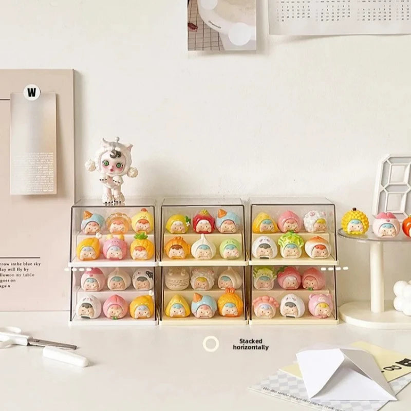 Six clear acrylic display boxes stacked horizontally on a tabletop, each holding multiple small pastel character figurines. The setup is styled in a clean, minimalist room with a cream wall, a small white character figurine on top, and desk accessories like paper and a pencil.