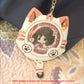 A cat-shaped acrylic keychain holder with a cartoon girl display insert, resting on a patterned quilt next to a dark navy bag. The keychain has orange paw-print accents, white cat ears, and a silver metal chain.