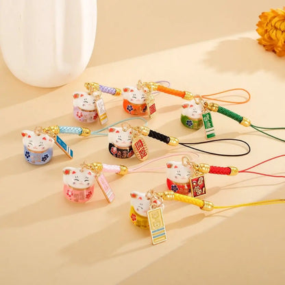 A set of colorful Japanese lucky cat (Maneki-neko) charm phone straps arranged on a beige surface with a white vase in the background.