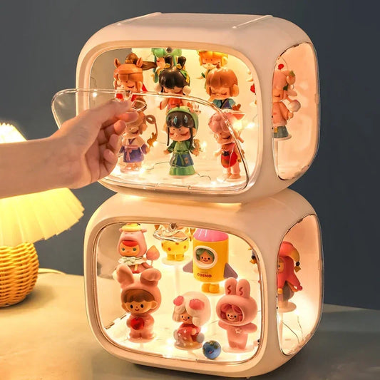 A hand is holding a clear acrylic panel in front of a stacked two-tier display case filled with small colorful collectible figurines. The case has rounded cube-shaped sides and warm white LED lighting inside, set on a tabletop with a lamp in the background.