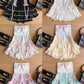 Six ruffled dresses arranged in a 2x3 grid on a wooden surface, each paired with a black handbag. The dresses come in color combinations of black, white, pink, yellow, lavender, and mint green, all featuring a pale green bodice with a pink bow and lace-up details.