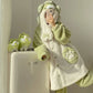 A person wearing a plush light green and off-white hooded robe with animal-inspired ear details and paw-print patches, taking a mirror selfie while leaning against a white appliance. Two matching plush accessories sit on the appliance beside them.