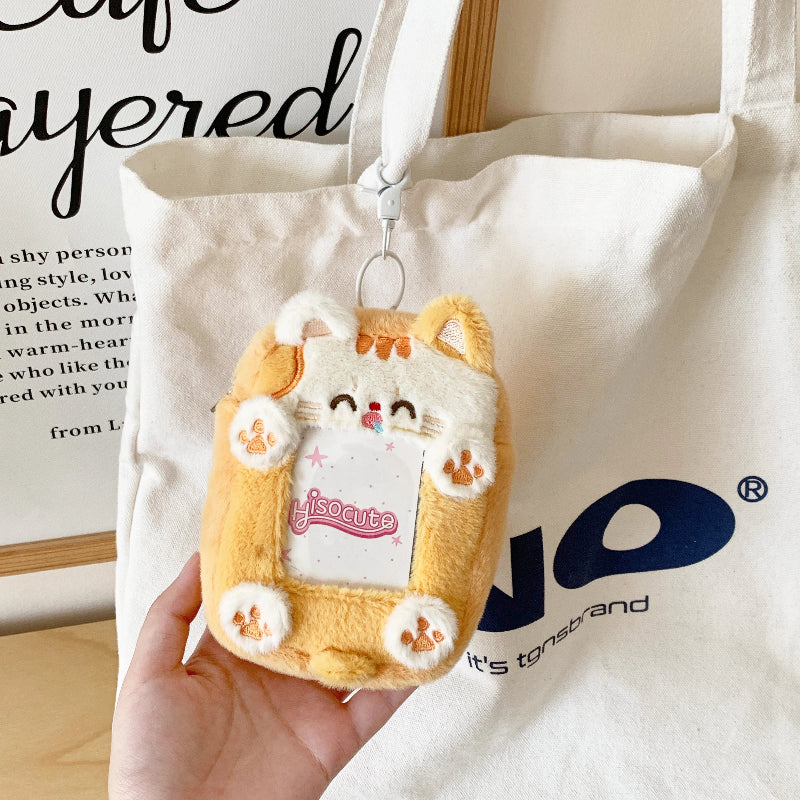 Plush keychain shaped like a cat in a bag held by a hand, with a white tote bag in the background.