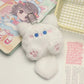 Plush keychain shaped like a white cat with pink accents on a surface with a card and basket in the background.