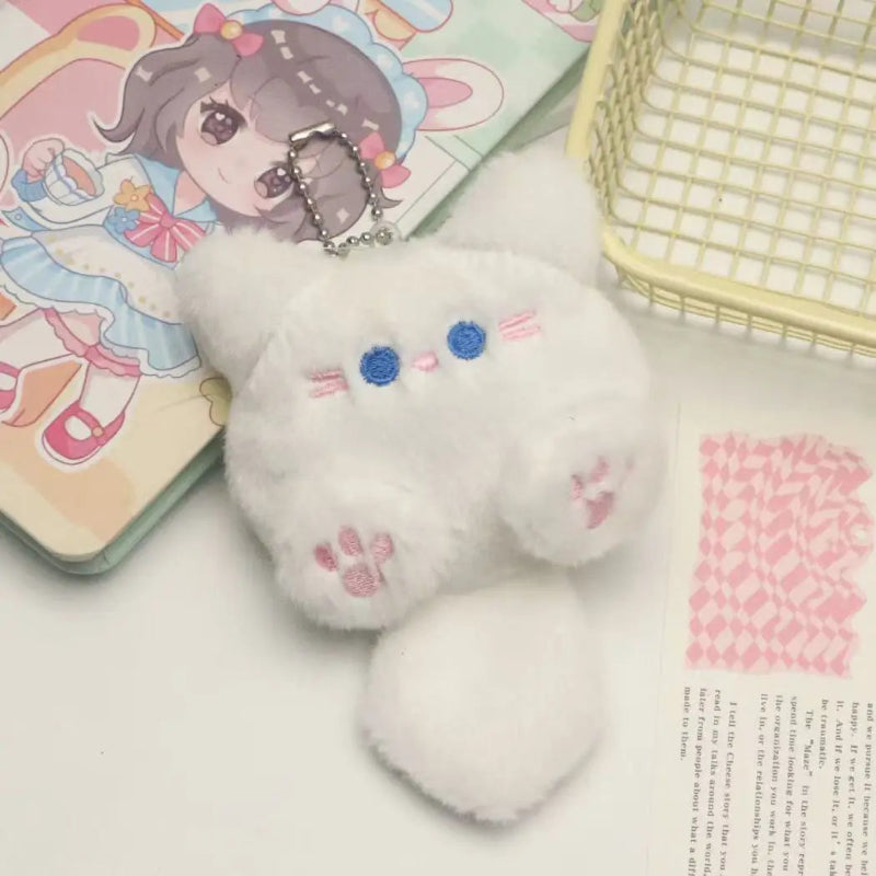 Plush keychain shaped like a white cat with pink accents on a surface with a card and basket in the background.