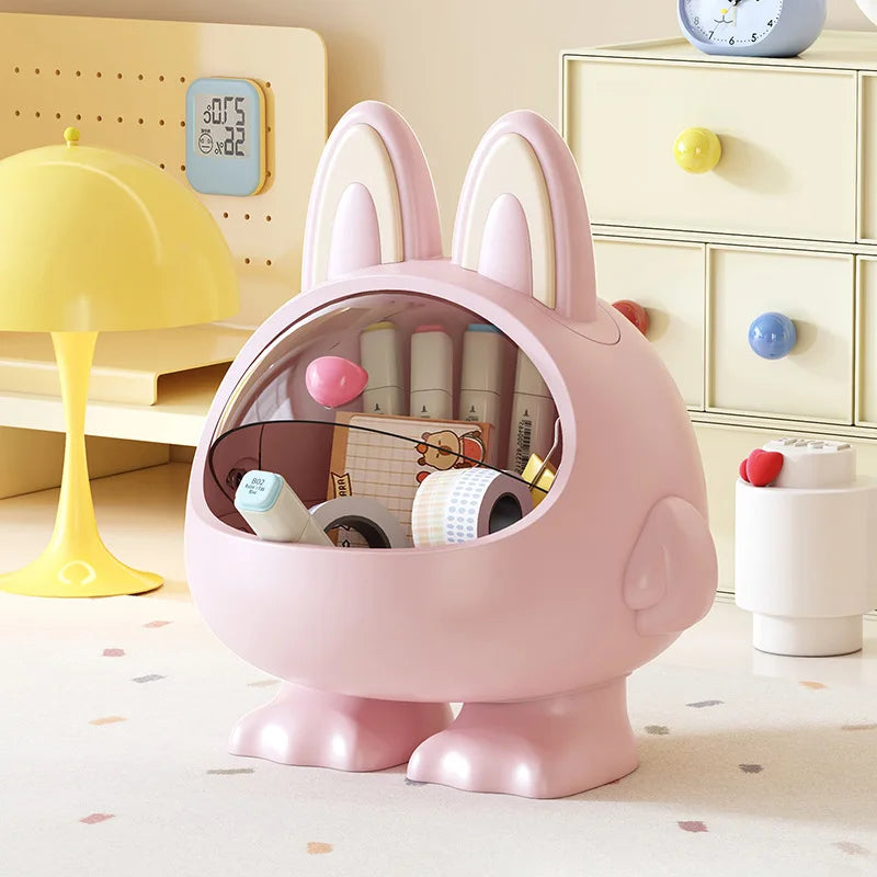A pink bunny-shaped desk organizer with long ears and a clear front window, filled with pens, sticky notes, and washi tape, set in a pastel-colored room with a yellow lamp and cream drawers.