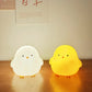 Two chick-shaped night lights, one white and one yellow, on a wooden surface.