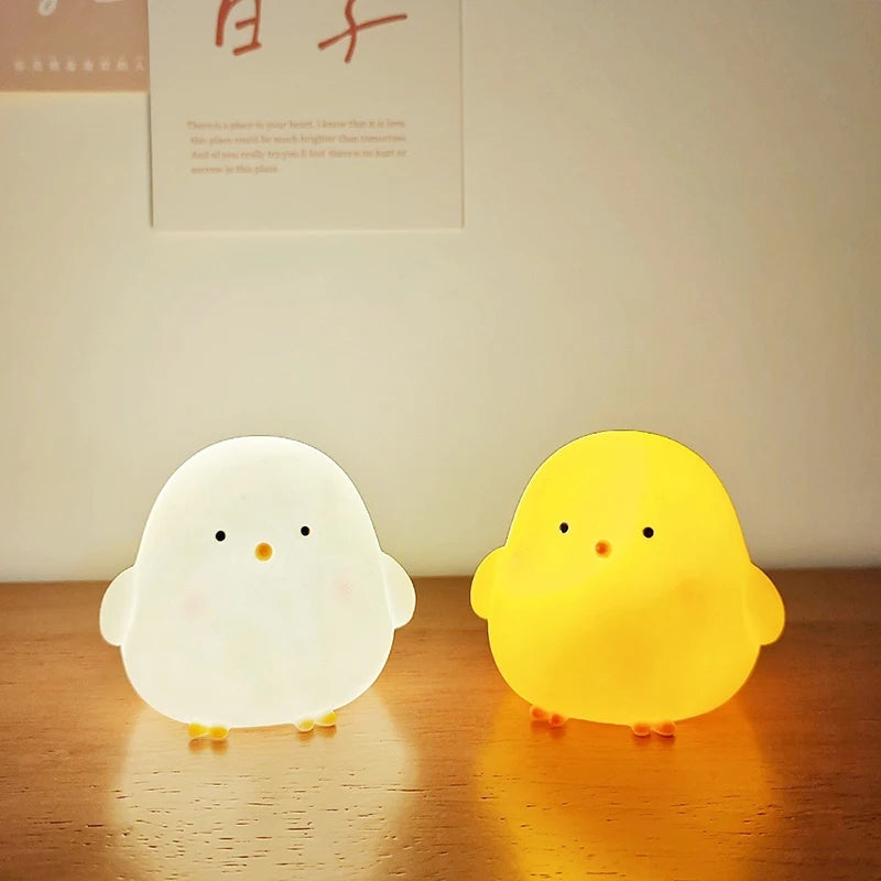 Two chick-shaped night lights, one white and one yellow, on a wooden surface.