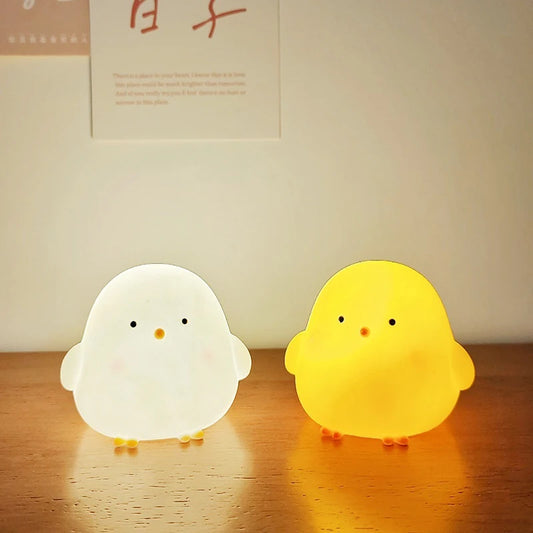 Two chick-shaped night lights, one white and one yellow, on a wooden surface.