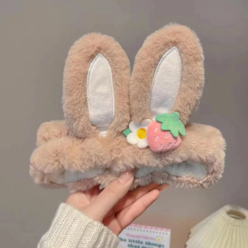 Fluffy kawaii bunny headbands with ears and a flower and strawberry decoration held by a hand against a neutral background.