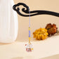 A small hanging charm with a purple braided cord, gold-toned metal accents, and a white cat figurine with red and gold details, displayed on a black decorative stand against a warm beige background.