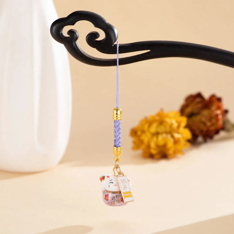 A small hanging charm with a purple braided cord, gold-toned metal accents, and a white cat figurine with red and gold details, displayed on a black decorative stand against a warm beige background.