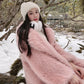 A woman wearing a fluffy light pink faux fur coat, a cream knit beanie, and matching cream ear muffs, posing outdoors in a snowy, mossy forest setting.
