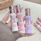 Three small lip glosses with crowns on lids held by a hand, against a neutral background.