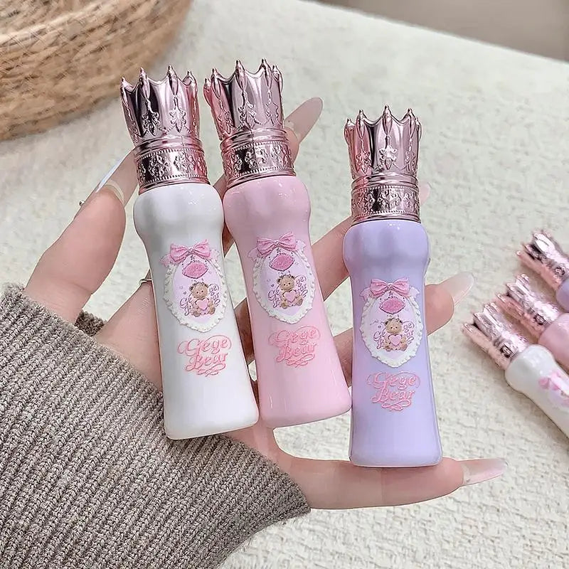Three small lip glosses with crowns on lids held by a hand, against a neutral background.