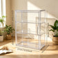 A clear acrylic three-tier storage cabinet with a side handle, shown in a sunlit room with plants and dimension lines and text indicating its measurements: 27cm/10.63inch wide, 36cm/14.