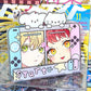 A novelty acrylic stand or plaque shaped like a retro handheld game console, featuring illustrated anime-style female characters on the left and right halves, with two white cloud shapes on top and the word 'Start' across the bottom. The product is photographed in a retail or desk setting with magazines and other items in the background.