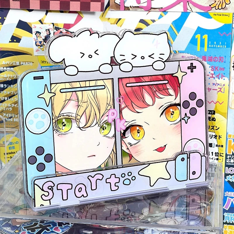 A novelty acrylic stand or plaque shaped like a retro handheld game console, featuring illustrated anime-style female characters on the left and right halves, with two white cloud shapes on top and the word 'Start' across the bottom. The product is photographed in a retail or desk setting with magazines and other items in the background.