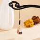 A small hanging charm with a black braided cord, gold-toned beads, and a decorative cat-shaped pendant, displayed against a warm beige background with a white vase and blurred decorative elements.