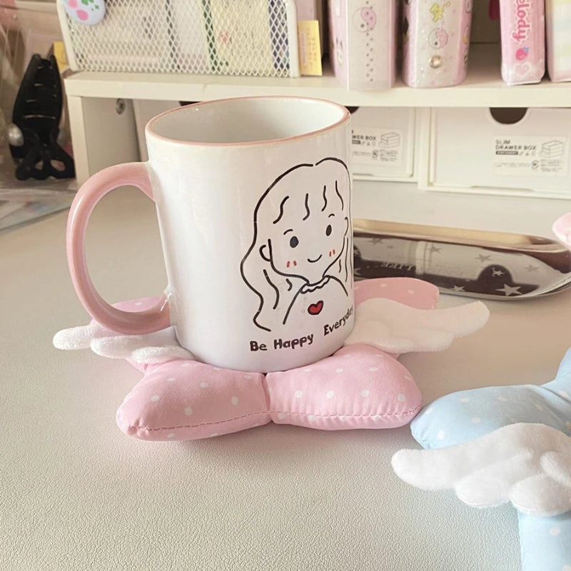 White mug with a cartoon character and 'Be Happy Everyday' text on a pink coaster with angel wings.