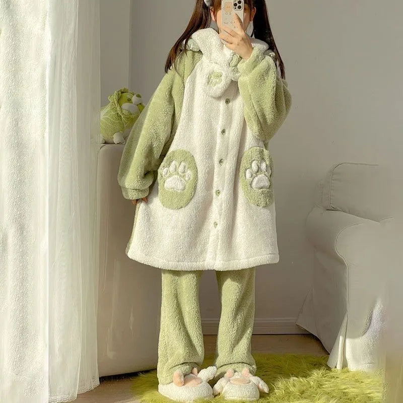 A person wearing a light green and off-white plush fleece pajama set with a hooded button-front top and matching pants, standing indoors near a window with a phone covering the face.