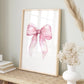 A framed art print featuring a watercolor pink ribbon bow on a white background, styled in a home decor setting on a woven table runner with dried grass and a white vase.