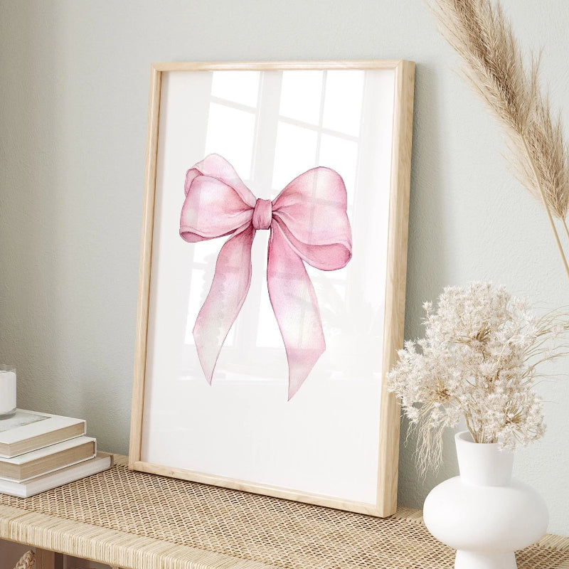 A framed art print featuring a watercolor pink ribbon bow on a white background, styled in a home decor setting on a woven table runner with dried grass and a white vase.