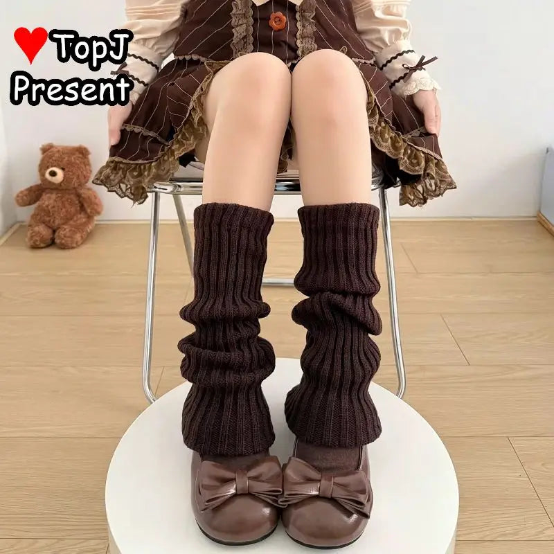 A person seated on a chair wearing dark brown ribbed leg warmers and matching brown bow shoes, with a dollhouse-style dress visible above the knees. A teddy bear sits on the floor in the background, and a 'TopJ Present' logo appears in the top left.